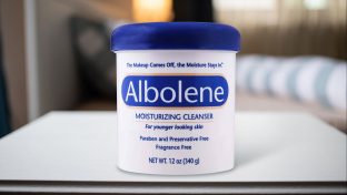 Photograph Of Albolene Container On White Table Top With Out Of Focus Bedding In Distant Background