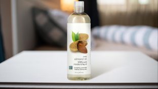 Photograph Of Almond Oil Bottle On White Table Top With Blurred Bedding In The Distant Background