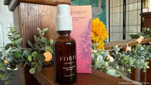 a bottle of foria awaken arousal oil with cbd and its box on a wood beam surrounded by greenery, florals, and lights, as the best cbd lube