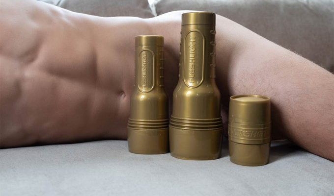 photograph of three fleshjack stamina training unit (stu) models in front of a nude man laying on a bed, the fleshjacks covering his groin