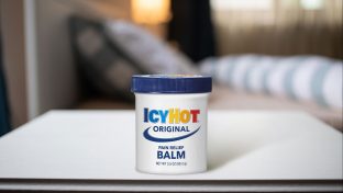 Photograph Of Icy Hot Container On White Tabletop With Out Of Focus Bedding In Distant Background