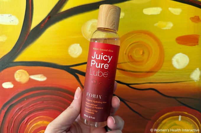 hand holding a bottle of foria juicy pure against a sunset painting with yellow, orange, and red paint strokes