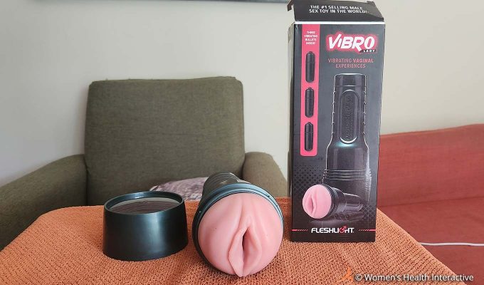 photograph of the fleshlight vibro lady with its cap removed and the vulva facing the viewer, laying on a cloth surface next to its black and red box