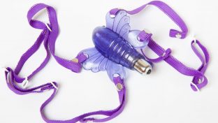 Closeup Photograph Of Purple Butterfly Vibrator With Matching Purple Harness Laying Against White Background