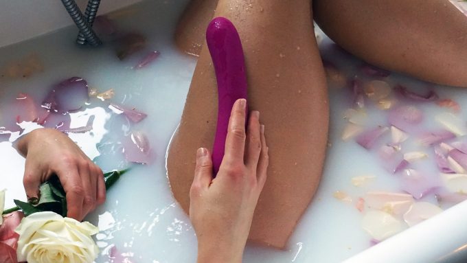 seductive photograph of a woman's leg in a milky bath filled with flower petals with a g-spot vibrator in her hand and resting against her thigh