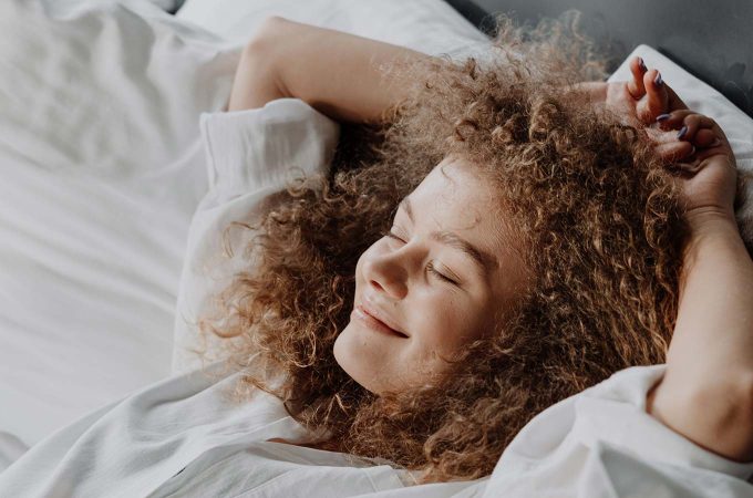 woman smiling with her eyes closed while laying back against pillows in bed